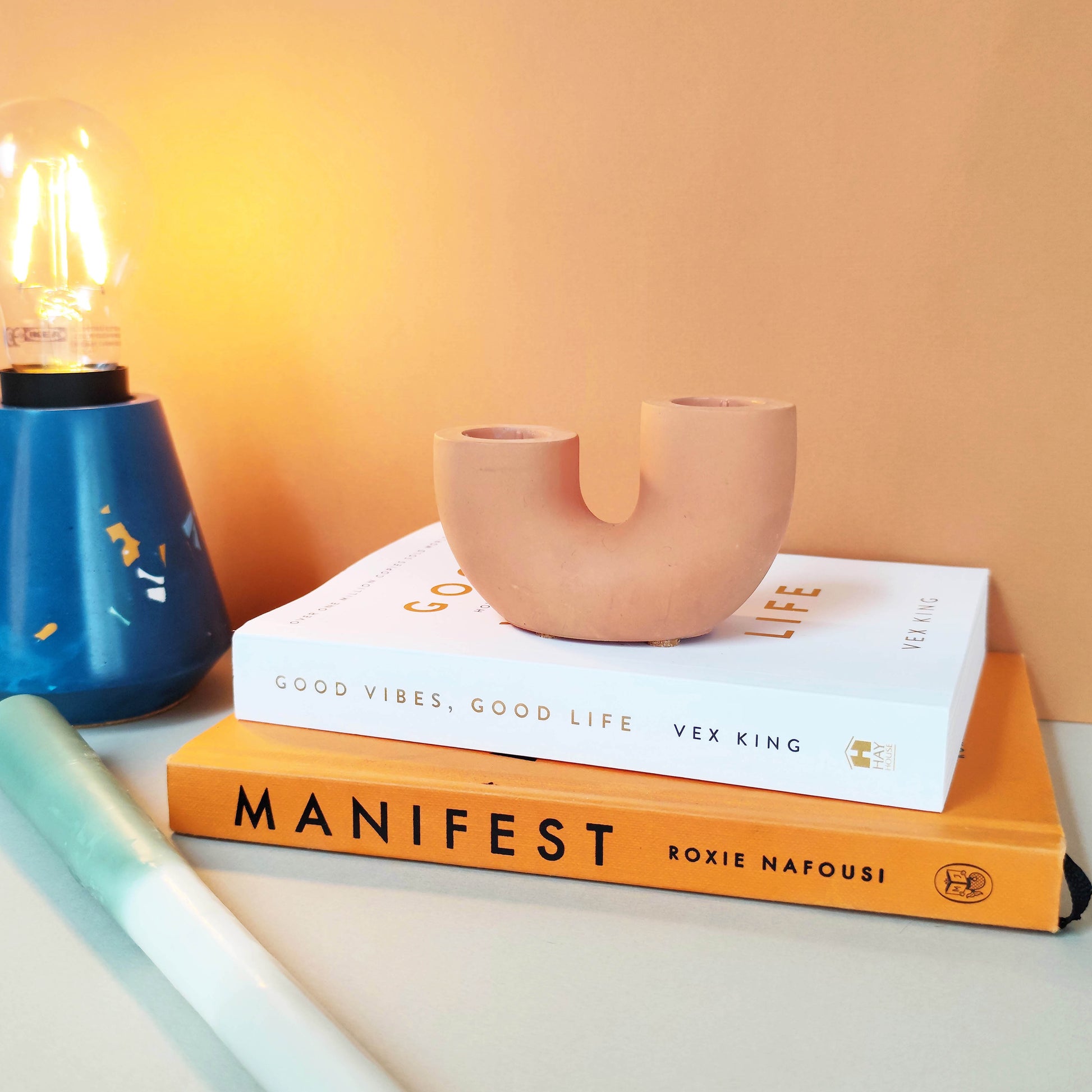 A dual U-shaped candle holder in a beige color, placed on top of two books titled 'Manifest' and 'Good Vibes, Good Life', next to a blue candle and a plant.