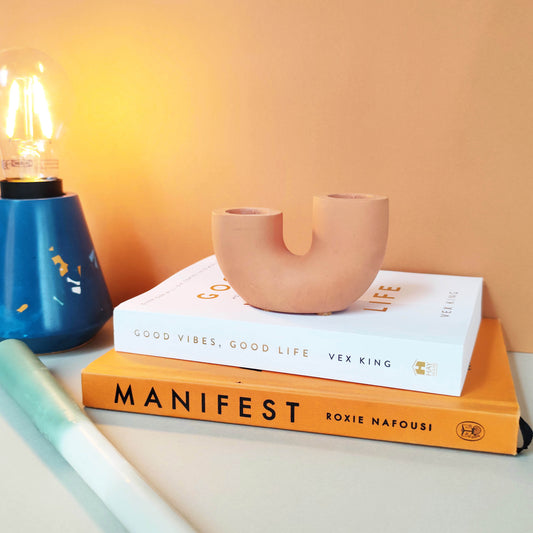 A dual U-shaped candle holder in a beige color, placed on top of two books titled 'Manifest' and 'Good Vibes, Good Life', next to a blue candle and a plant.