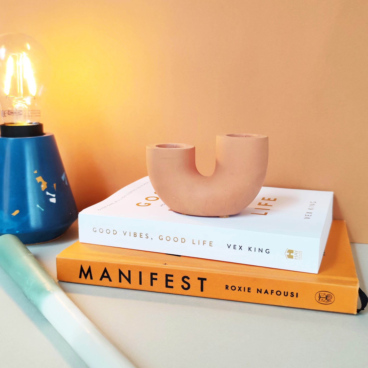 A dual U-shaped candle holder in a beige color, placed on top of two books titled 'Manifest' and 'Good Vibes, Good Life', next to a blue candle and a plant.