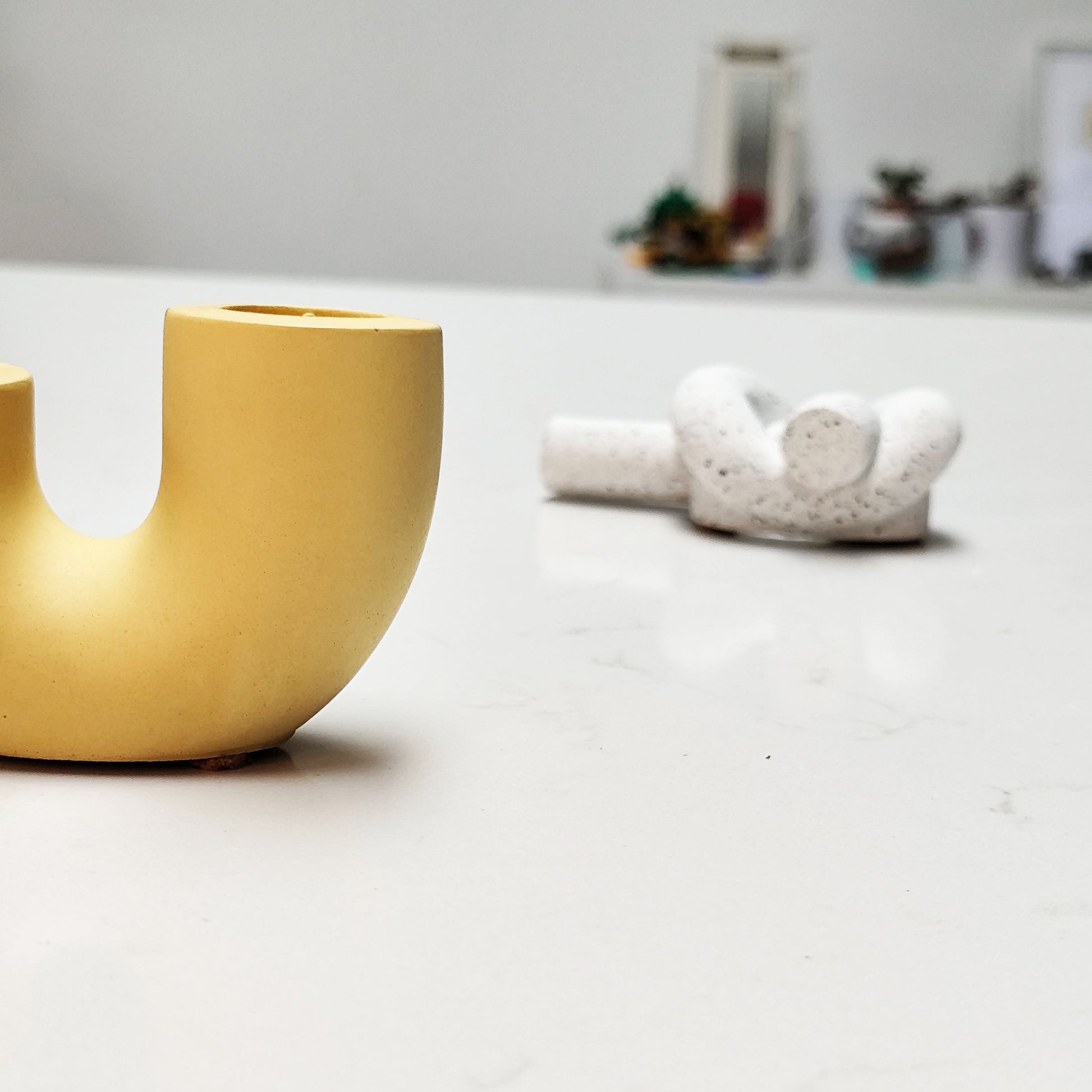 A dual U-shaped candle holder in a bright yellow color, placed on kitchen island with twist candle holder in background