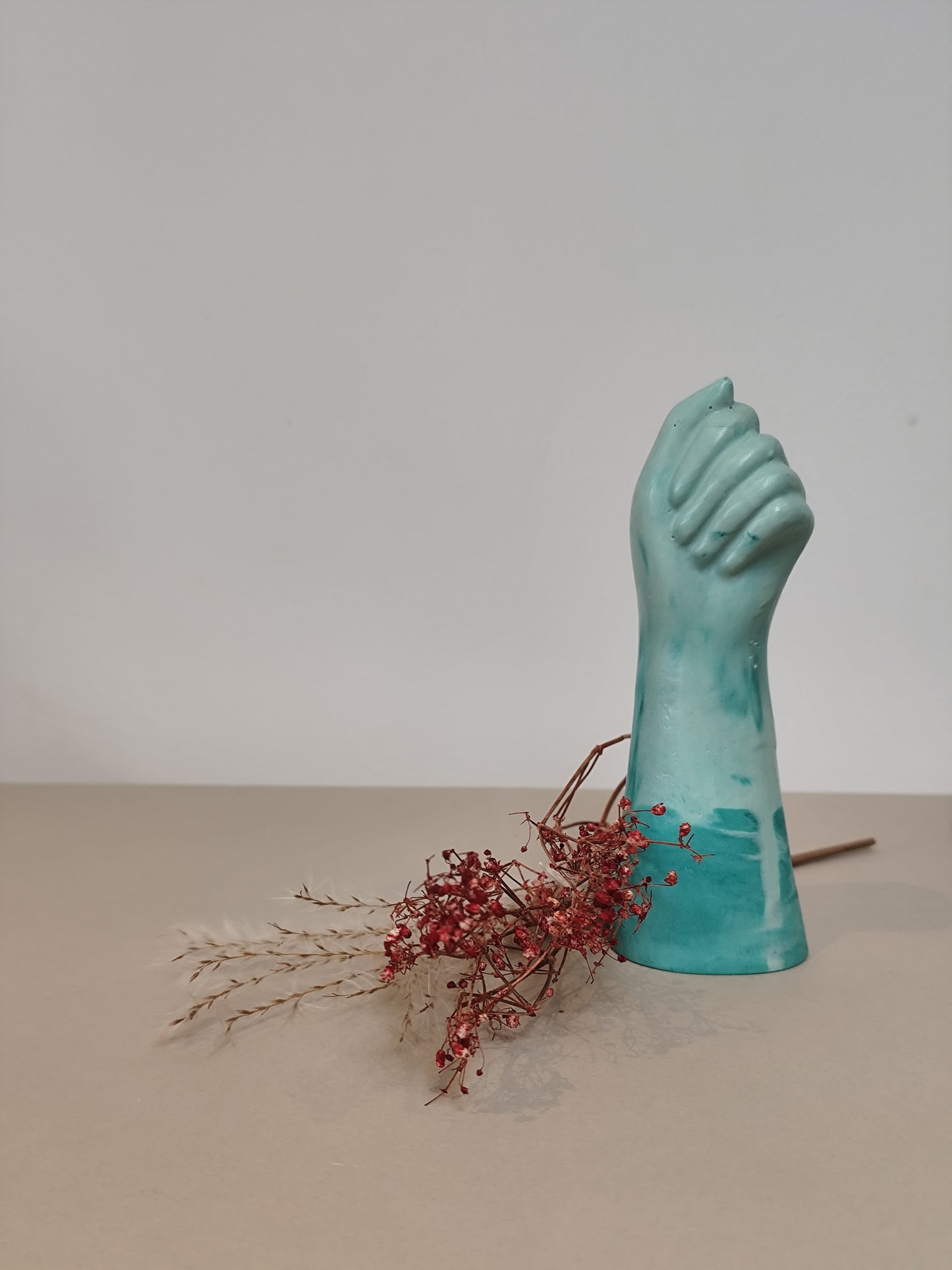 Turquoise handmade vase shaped like a fist holding red berries on a beige surface with a white background