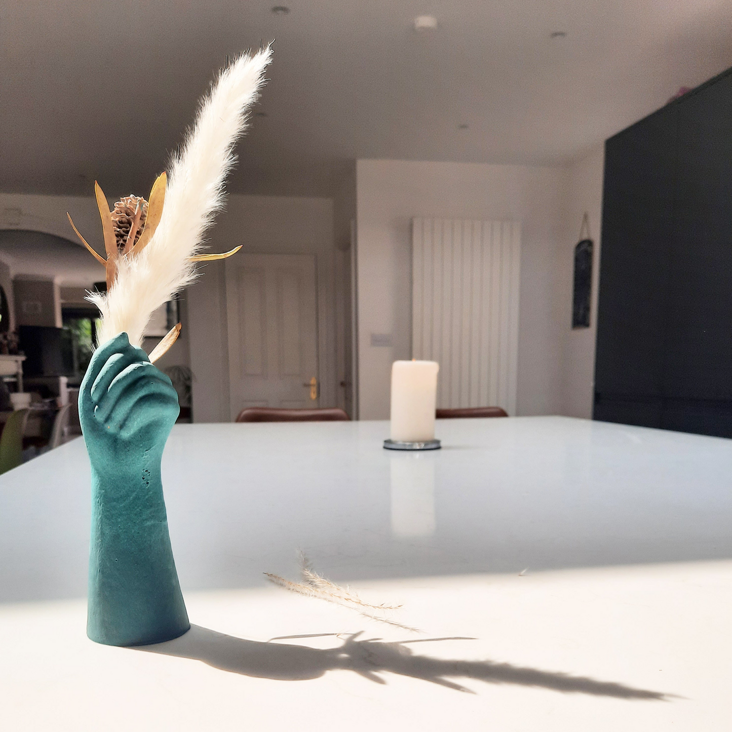 A unique, handcrafted vase with a marbling effect, in a green color, in the shape of a closed fist, placed on a kitchen countertop with dried flowers on top.