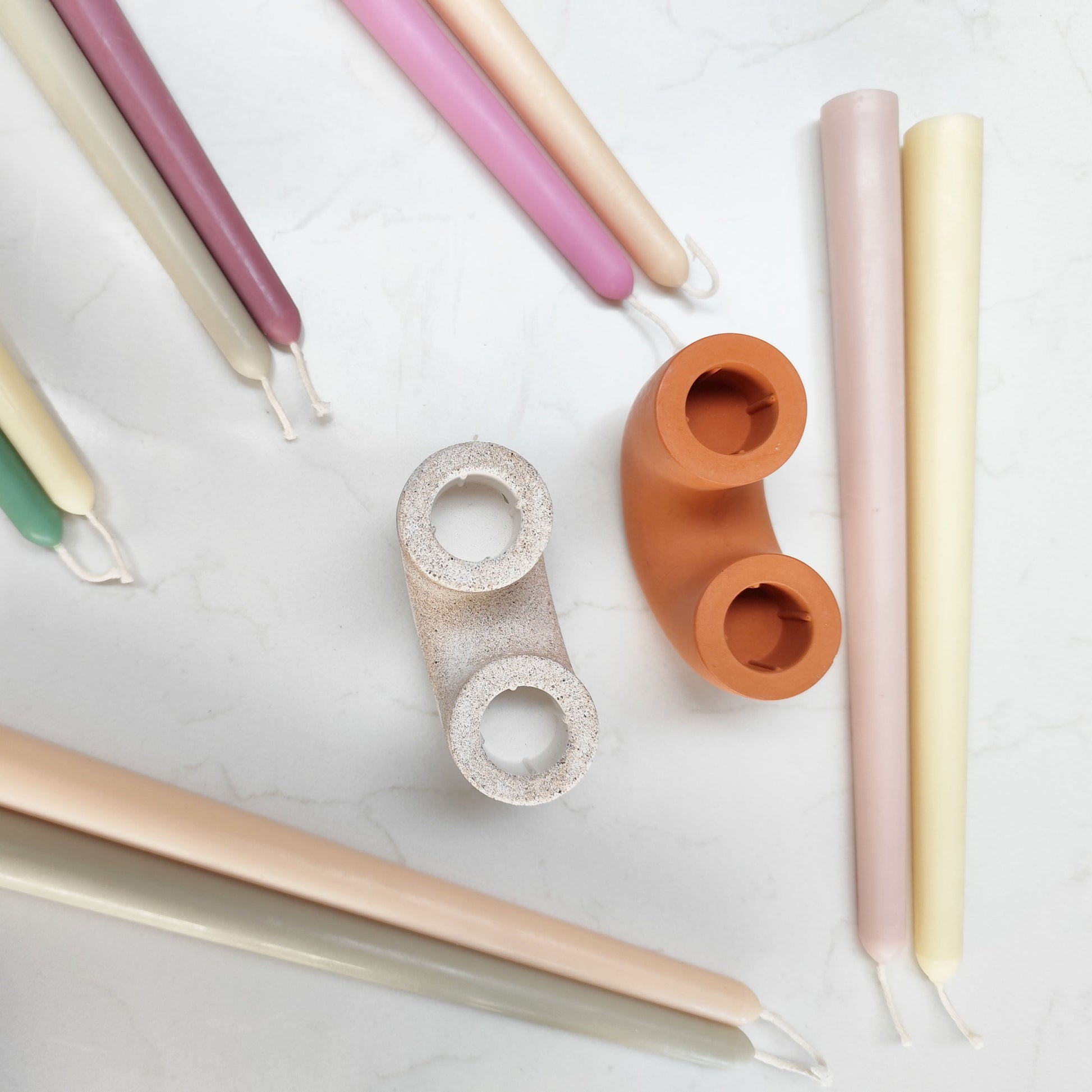 Two candle holders white and Terracotta_colour surrounded by pairs of natural beeswax dinner candles in varitey of pastel colours