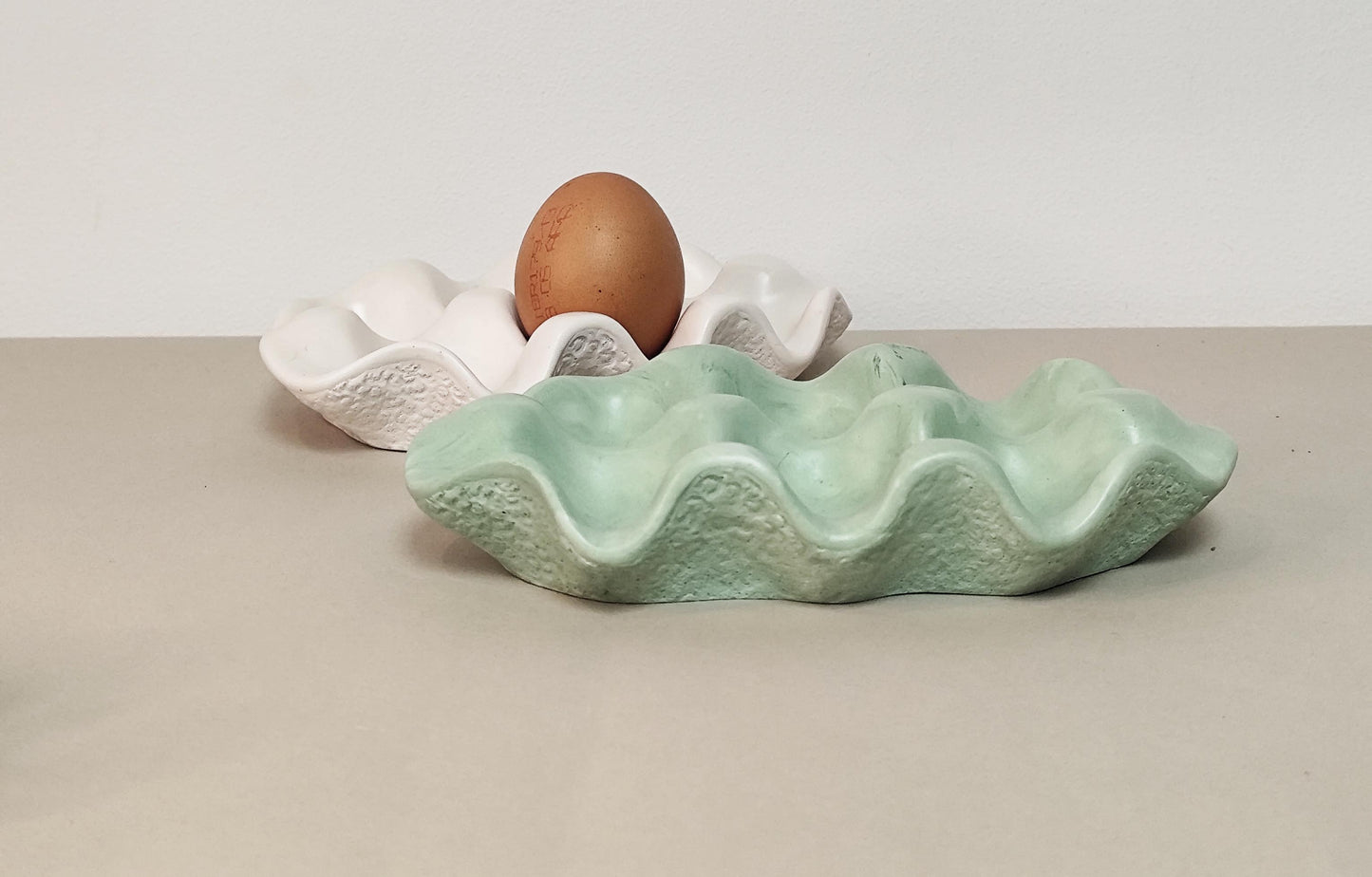 Jesmonite handmade egg holder shaped like a shell, in a sage green with a brown egg on a beige surface.