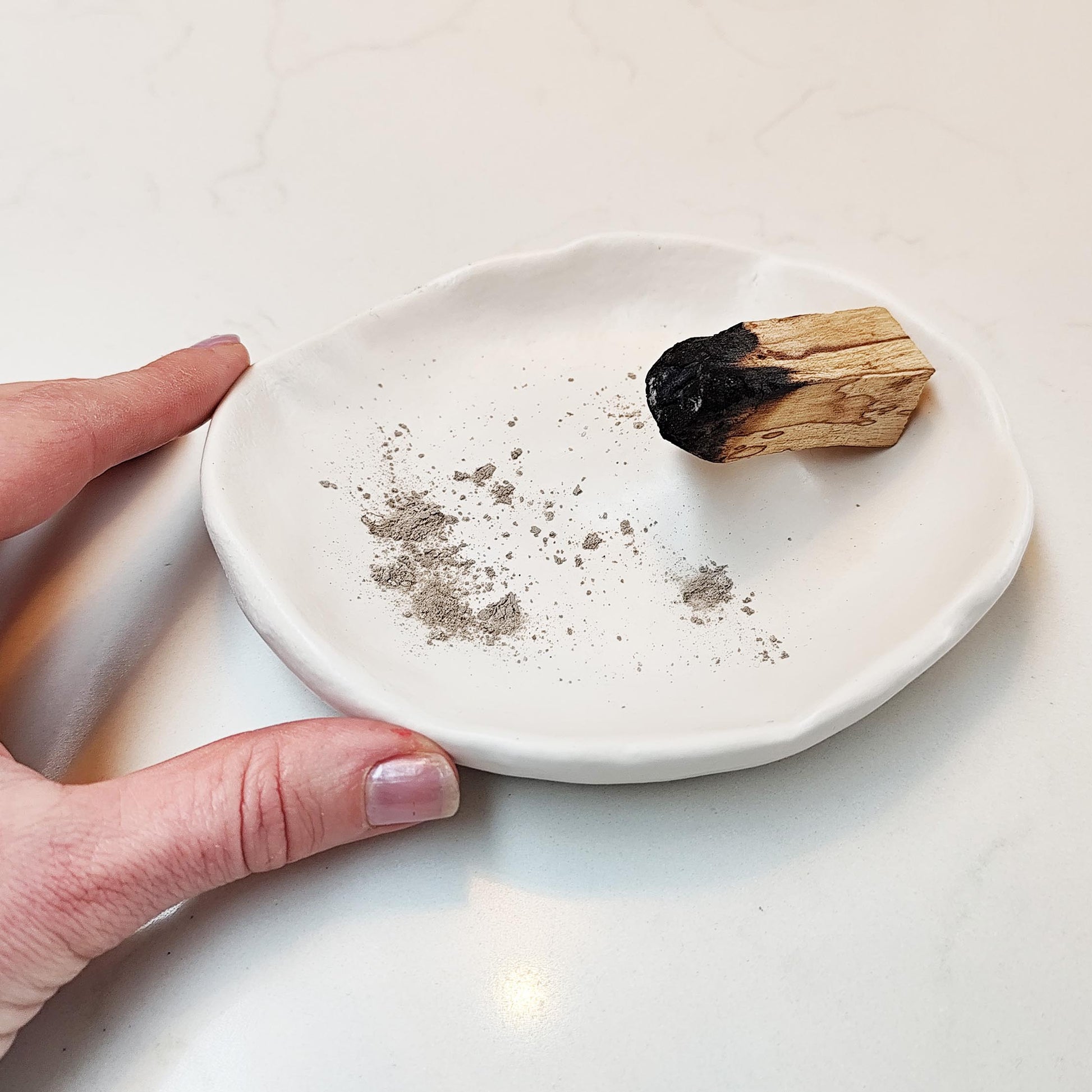 Hand holding a white burner with incense ashes and a piece of burned wood on a light background
